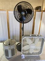 Front view of all three fans: Lasko black pedestal oscillating fan, Lasko white box fan, Duracraft HEPA filter fan