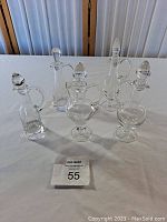 Five glass oil and vinegar bottles with matching glass stoppers arranged on a white tablecloth. All bottles are clear and approximately 5.5 inches tall, each with a handle and narrow spout.