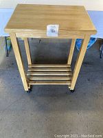 Photo of light-colored solid wood kitchen cart with flat top surface and slatted lower shelf, caster wheels on legs nearby.