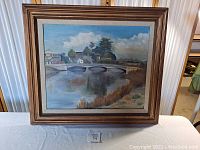 Front view of the traditional oil painting "San Lorenzo River" by Ray Lewis in wooden frame on a white table.