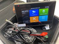 HD CCTV tester device with screen active showing menu, connected cables visible with a DC power plug and alligator clips, inside a fabric carrying case.
