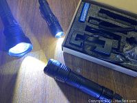 Four black flashlights on a wooden surface, two turned on emitting white light, with accessories such as charger and batteries in foam insert box.