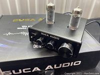 Front-side view of the Suca Audio tube headphone amplifier showing the two tubes on top and control knobs including power, tube bass, and volume knob.