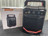 Photo showing Socovez travel speaker beside its original box, revealing speaker front with handle, control knobs, USB ports, and aux input.