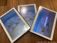 Three tablets laid out on wood surface with screens on, showing home screens and time.