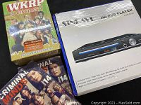 Photo of WKRP in Cincinnati DVD box set, two Criminal Minds Season 14 DVD box sets, and Sylvania HD DVD player box with player and remote on table.