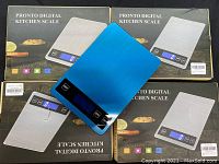 Four Pronto digital kitchen scales visible with three in boxes and one outside showing blue brushed surface and digital screen.