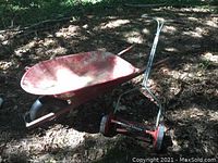 Wheelbarrow and push mower displayed outdoors on dirt and leaves under shade