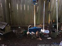 Wide view of garden tools leaning against wooden fence including rakes, shovels, large jug, decorative bird houses, metal watering can.