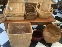 Photo showing assorted woven baskets including large hamper, smaller baskets and wooden trays on a wooden table and floor with black and white tiled floor.