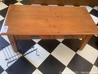 Top and front angle of wood coffee table with two drawers and adjacent metal magazine rack on checkerboard tile floor.