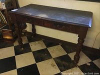 Full view of the vintage wooden desk showing two drawers and turned legs, placed on checkered floor.