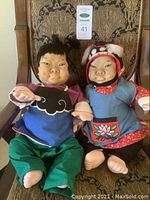 Two dolls seated on a chair showing full front view, detailed facial features, and traditional clothing.