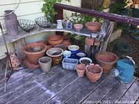 Wide angle showing multiple terra cotta and ceramic pots, metal watering can, wire hanging planters on wooden deck