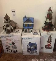 Photo of three lighted decorative houses on their boxes: church on left, house in center, lighthouse on right