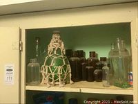 Shelf of assorted vintage apothecary jars including the large green bottle with macrame netting and various amber and clear bottles.