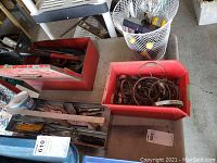 Photo showing red metal tool box with hand tools, red plastic bin containing metal hose clamps, utility knives, and nearby metal bathroom waste can.