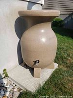 Full front view of beige urn-shaped rain barrel positioned outdoors showing block feet and tap.