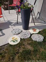 Photo showing four round garden stepping stones and a tall woven-style black planter with a live flowering plant.