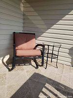 Patio rocker with rust-colored cushions next to black metal side table with a glass top, placed against a beige exterior wall and patterned outdoor rug.