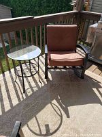 Patio rocker with rust-colored cushions next to a round glass top metal side table on an outdoor deck area.