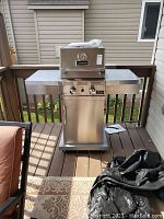 Full view of the stainless steel outdoor gas BBQ with side shelves and wheeled base on a wooden deck.
