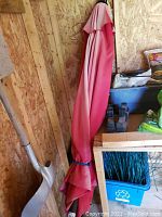Closed pink patio umbrella leaning against wooden wall inside a shed, showing sun discoloration on fabric.