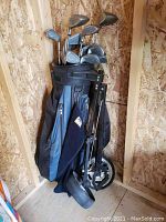 Photo showing golf clubs in blue and black golf bag with attached black golf cart in a wooden shed corner.