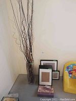 Photo of glass vase with tall decorative sticks and several black frames stacked against wall near yellow child's chair.