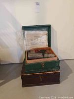 Green wooden box containing various metal Meccano construction parts including gears, wheels, and plates.