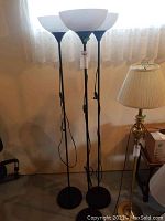 Three black metal floor lamps standing together in front of a window with lace curtains. Each has a white glass bowl-shaped shade and round weighted base.