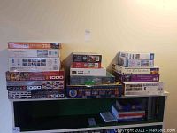 Lot of assorted puzzle boxes stacked on a black shelf, showing puzzle brands, piece counts, and various images on box art.