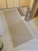 Full view of longer beige and brown geometric pattern area rug on floor with smaller striped rugs and two rolled rugs nearby.