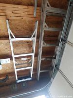 Two aluminum ladders leaning against a wall in a wooden garage. One is a 3-step ladder with a blue top cap, the other is a taller straight ladder approx. 5 feet.