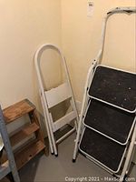 Three folding stepladders in a corner: one wooden, two white metal with black steps.