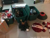 Photo showing green plastic Christmas tree stand, oversized candy cane ornaments, 'Merry Christmas' wreath decoration, plus multiple green storage bins filled with various Christmas decorations and lights.