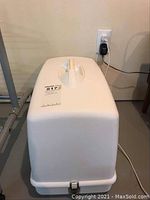 White plastic carrying case for the sewing machine, closed with a handle on top and latch at the front.