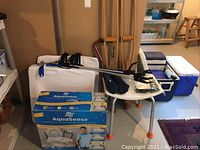 Health aids including white shower chair, Aqua Sense raised toilet seat in box, wooden crutches, cane, black stethoscope, slippers, shoe grippers.