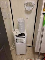 Haier portable air conditioner with flexible exhaust pipe and plexiglass panel shown. Unit is positioned upright against the wall with pipe and vent attachment on top.