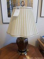 Full view of the lamp on a wooden table next to framed pictures and books. Shows the brass base and pleated cream fabric shade with scalloped edge.