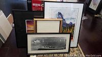 Four framed artworks shown leaning against a wall on the floor, including the black and white ship photo, colorful mountain landscape, sunset seascape, and empty frame.