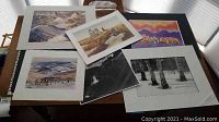 Six unframed art prints and photography pieces displayed on a table, showing winter landscapes, a lighthouse, a black and white forest photo, and an abstract colorful cityscape.