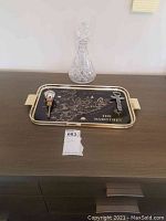 Full lot view showing the cut glass decanter, the pewter bottle opener with cork pour spout, and the black and gold serving tray with map design on a wooden surface.