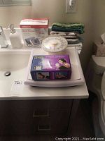 Photo showing all three main items: Sunbeam bath scale, Omron blood pressure monitor box, Dr Ho's Muscle Therapy box stacked on white surface with folded towels in background.