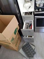Kitchen drawers open showing assorted cooking utensils such as spoons, spatulas, and whisks, along with muffin pans and baking sheet placed on the floor.