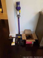 Full view of stick vacuum standing with wand attached plus box of attachments and charger on floor