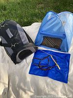 Photo of black and gray soft-sided cat travel carrier, blue pop-up cat tent, blue cooling mat, and multi-colored leash and collar set on white cloth on grass.