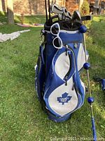 Blue and white Toronto Maple Leafs golf bag with multiple zippered compartments, carrying handle, and shoulder strap; white sunglasses hanging on bag.