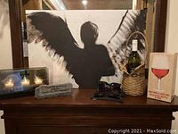 Framed angel silhouette print, candle holder with lit candles, wine basket, large wine glass decoration, stone sign, and three pairs of sunglasses on wooden dresser.