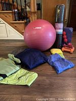 Photo showing the pink 18 inch exercise ball labeled 'The FIRM', two yoga mats (black and purple), folded yellow stretch band, 1 lb and 3 lb dumbbells in orange, red massager, and workout clothing including blue sweatshirt and green top.